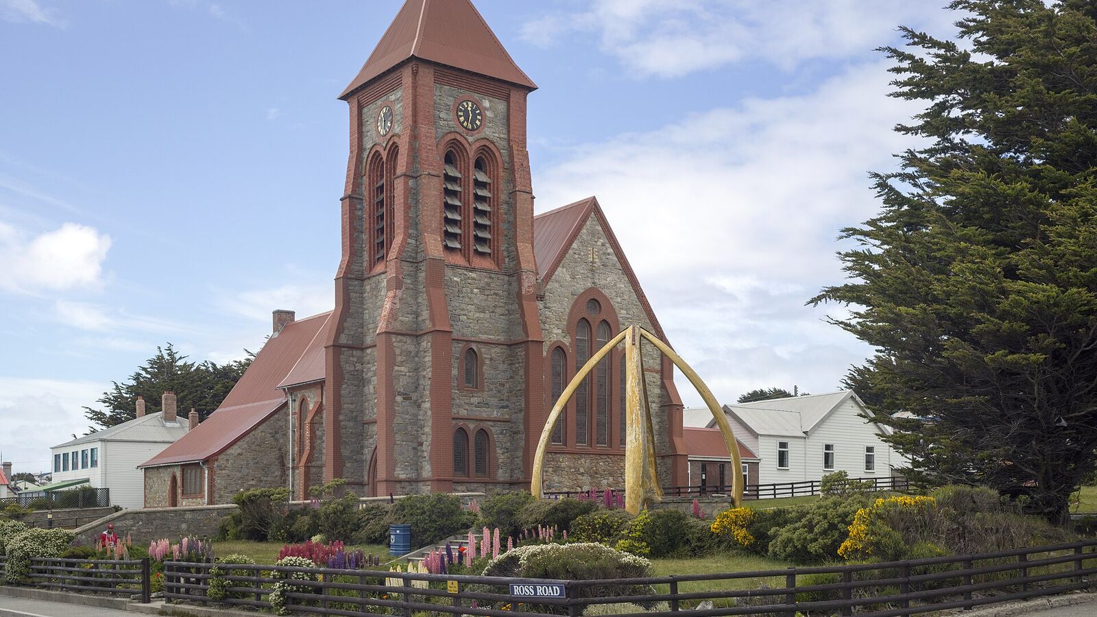 De bakstenen Christ Church Cathedral in Stanley met de boog van walvisbotten ervoor, aan een straat met bloemen