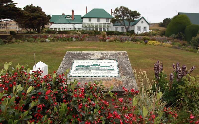 Government House in Stanley, een wit koloniaal huis met groen dak achter een tuin vol rode bloemen en een gedenksteen