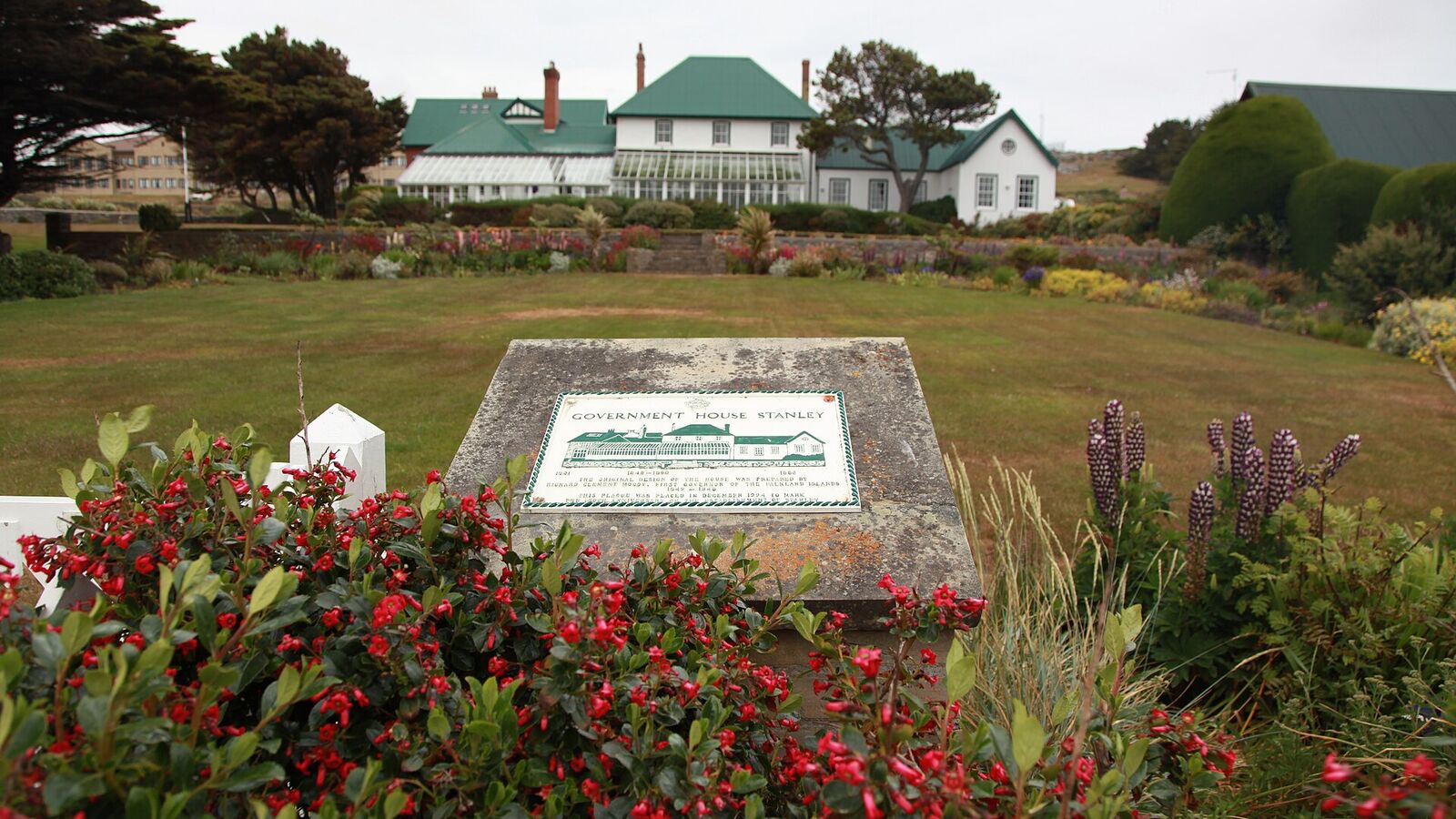 Government House in Stanley, een wit koloniaal huis met groen dak achter een tuin vol rode bloemen en een gedenksteen