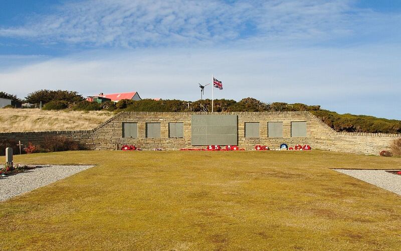 De ronde Britse oorlogsbegraafplaats van San Carlos met een stenen gedenkmuur, kruisen en een vlaggenmast onder een blauwe lucht
